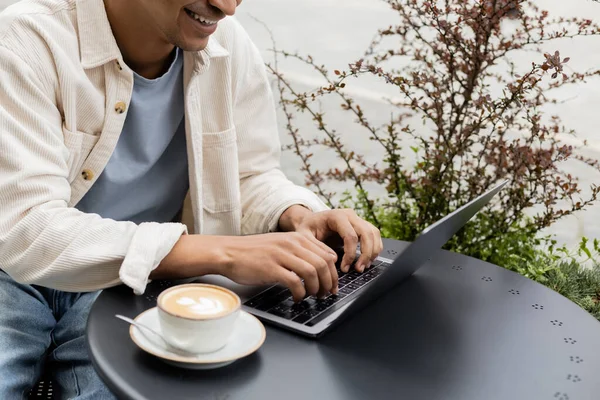 Cropped view of happy african american man using laptop near cup of latte on table in cafe terrace — Stock Photo