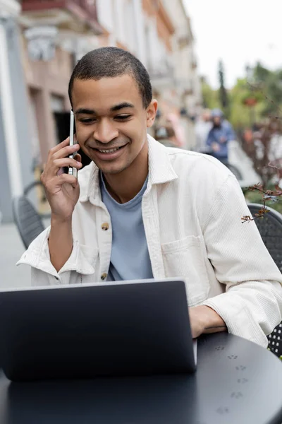 Freelancer afroamericano feliz hablando en smartphone cerca del portátil en la terraza de la cafetería - foto de stock