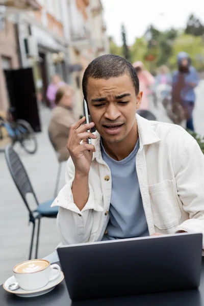 Young african american freelancer talking on smartphone near laptop on cafe terrace — Stock Photo