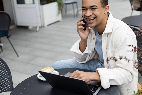 Alegre afroamericano freelancer hablando en smartphone cerca del portátil en la terraza de la cafetería - foto de stock