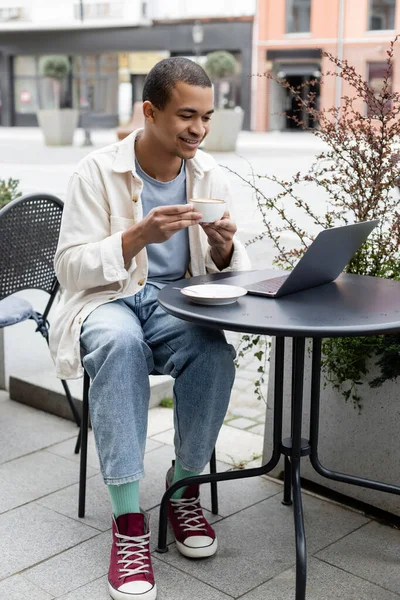 Cheerful african american freelancer drinking coffee and using laptop on cafe terrace — Stock Photo