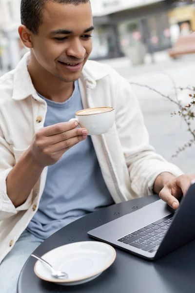 Sonriente freelancer afroamericano sosteniendo taza de café y utilizando el ordenador portátil en la terraza de la cafetería - foto de stock