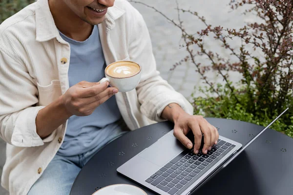 Обрезанный вид улыбающегося молодого африканского фрилансера, пьющего кофе и пользующегося ноутбуком на террасе кафе — стоковое фото