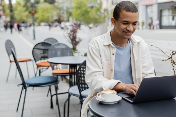 Sonriente freelancer afroamericano usando laptop cerca de capuchino en la terraza de la cafetería - foto de stock