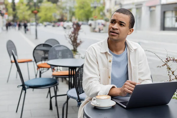 Рассеянный африканский американский фрилансер смотрит вдаль возле ноутбука и чашки капучино на террасе кафе — стоковое фото