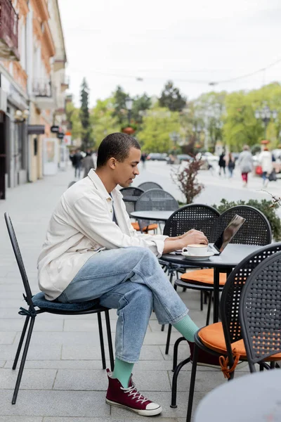 Vista lateral del joven afroamericano freelancer trabajando remotamente en el ordenador portátil cerca de la taza de capuchino en la terraza de la cafetería - foto de stock