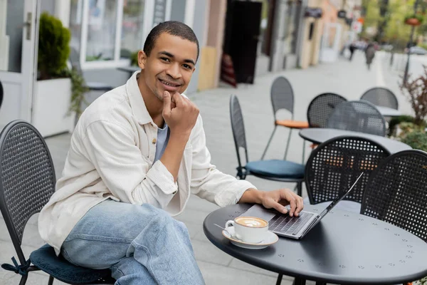 Улыбающийся африканский фрилансер смотрит в камеру рядом с ноутбуком и чашкой капучино на террасе кафе — стоковое фото