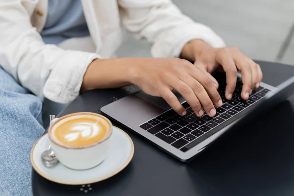 Vista recortada de freelancer afroamericano trabajando remotamente en el portátil cerca de la taza de capuchino en la terraza de la cafetería - foto de stock