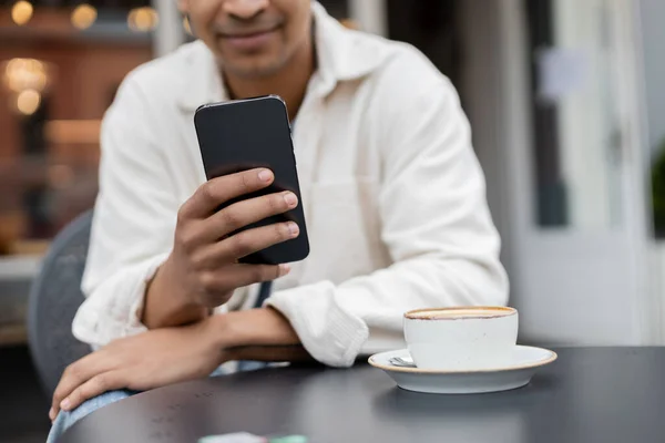 Vue recadrée de l'homme afro-américain flou texter sur smartphone près de tasse de cappuccino sur la table sur la terrasse du café — Photo de stock