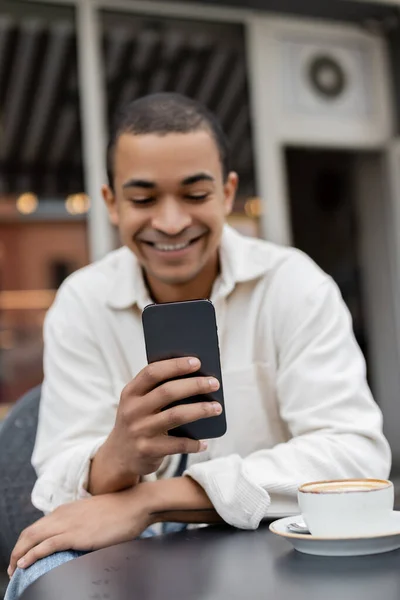 Homme afro-américain joyeux textos sur smartphone près de tasse de cappuccino sur la table sur la terrasse du café — Photo de stock