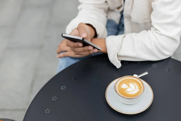 Vista parcial del hombre afroamericano mensajes de texto en el teléfono inteligente cerca de la taza de capuchino en la mesa - foto de stock