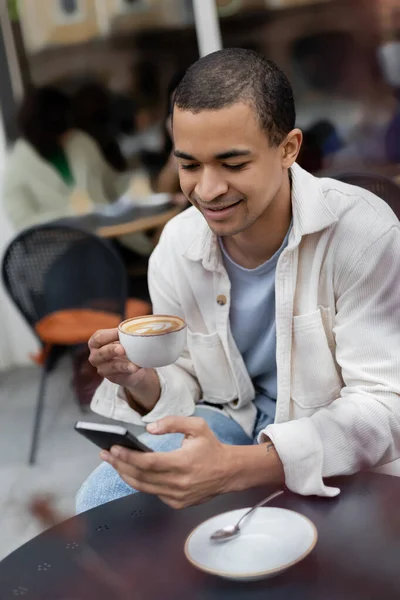 Улыбающийся африканский американец пишет смс на смартфоне и держит чашку капучино на террасе кафе — стоковое фото