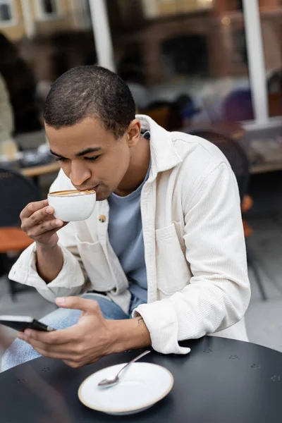 Hombre afroamericano mensajes de texto en el teléfono inteligente y beber capuchino en la terraza de la cafetería - foto de stock