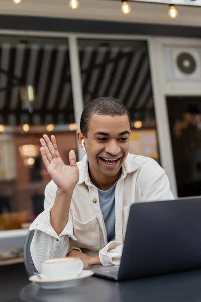 Счастливый африканский американец мужчина машет рукой во время видеозвонка на ноутбуке на летней террасе в кафе — стоковое фото