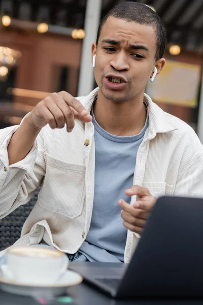 Afrikanisch-amerikanischer Freiberufler gestikuliert mit drahtlosen Kopfhörern während eines Videogesprächs auf der Sommerterrasse — Stockfoto