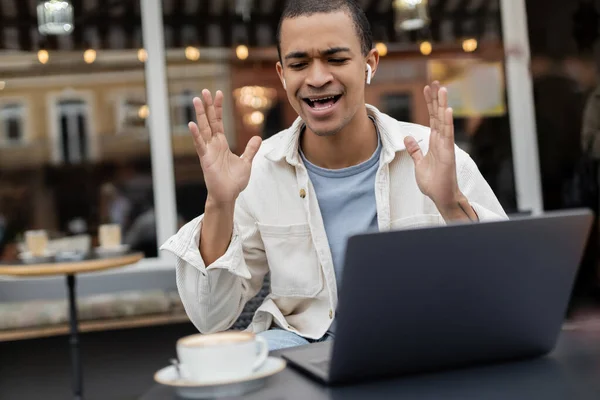 Sorprendido afroamericano freelancer en auriculares inalámbricos gesto durante la videollamada en la terraza de la cafetería - foto de stock