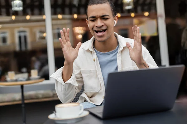 Shocked african american freelancer in wireless earphones gesturing during video call in cafe terrace — Stock Photo