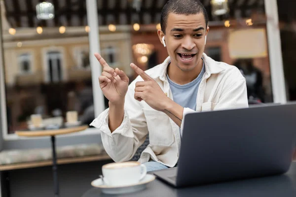 Freelancer afroamericano en auriculares inalámbricos apuntando a distancia durante una videollamada en la terraza de la cafetería - foto de stock