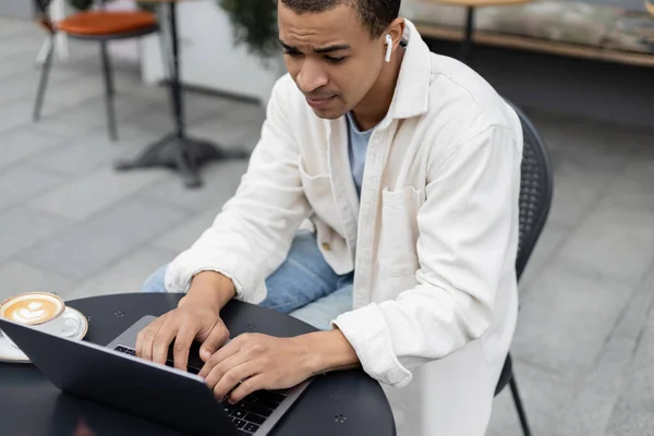 African american freelancer in wireless earphones using laptop near cappuccino on summer terrace — Stock Photo