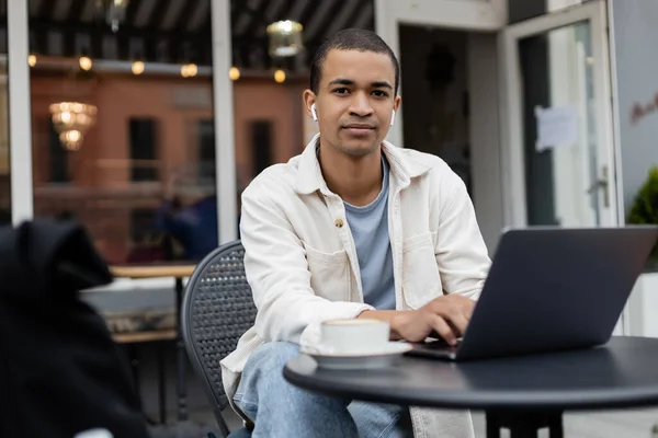 Freelance afroamericano in auricolari senza fili seduto vicino a tazza di caffè e laptop sulla terrazza estiva — Foto stock