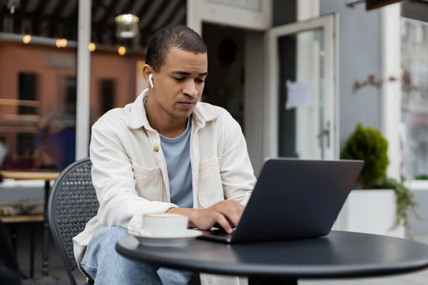 Freelancer afroamericano en auriculares inalámbricos sentado cerca de la taza de café y el uso de la computadora portátil en la terraza de verano - foto de stock