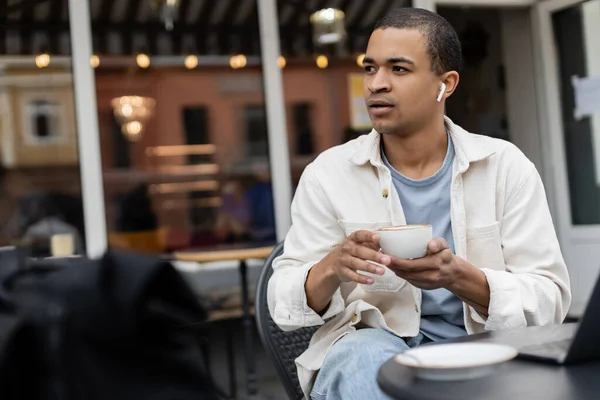 Афроамериканський чоловік у бездротових навушниках тримає чашку, сидячи біля ноутбука на літній терасі — стокове фото