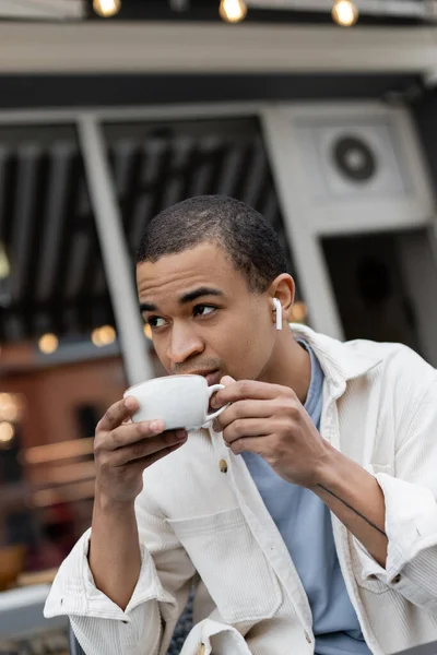 African american man in wireless earphones drinking coffee on summer terrace — Stock Photo