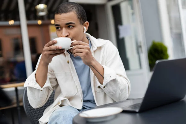 Африканский американский фрилансер в беспроводных наушниках пьет кофе возле ноутбука на летней террасе — стоковое фото
