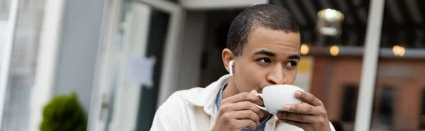 Afro-américain dans les écouteurs sans fil boire du café sur la terrasse d'été, bannière — Photo de stock