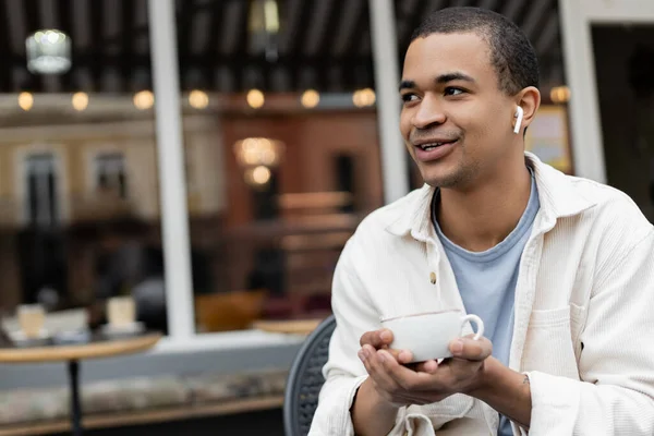 Happy african american man in wireless earphones holding cup of coffee on summer terrace — Stock Photo