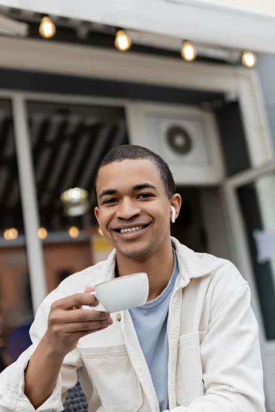 Gioioso uomo africano americano in auricolari senza fili che tengono una tazza di caffè sulla terrazza estiva — Foto stock