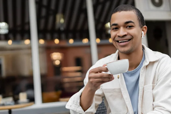 Cheerful african american man in wireless earphones holding cup of coffee on summer terrace — Stock Photo