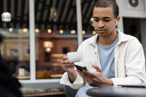 African american man in wireless earphones looking at smartphone while holding cup of coffee on summer terrace — Stock Photo