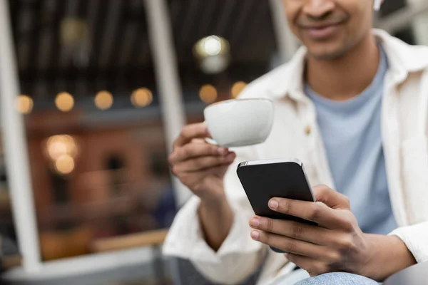 Vista ritagliata di felice uomo africano americano in auricolari wireless utilizzando smartphone mentre si tiene in mano una tazza di caffè — Foto stock