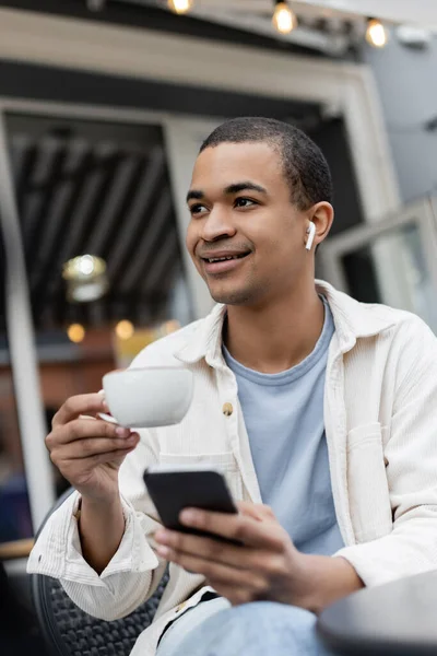 Uomo afro-americano sorridente con auricolari senza fili che reggono una tazza di caffè e smartphone sulla terrazza estiva — Foto stock