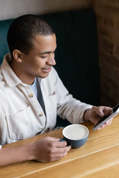 Sonriente y joven afroamericano hombre usando teléfono inteligente y la celebración de capuchino en la cafetería - foto de stock