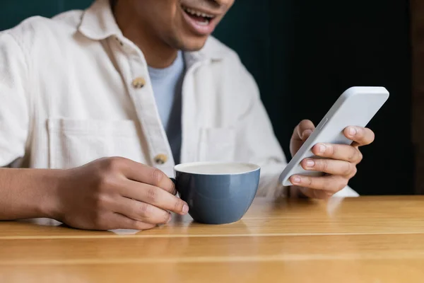 Vista recortada de alegre hombre afroamericano mensajes de texto en el teléfono inteligente cerca de la taza en la mesa - foto de stock