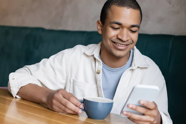 Sorridente e giovane afroamericano messaggistica su smartphone in caffetteria — Foto stock