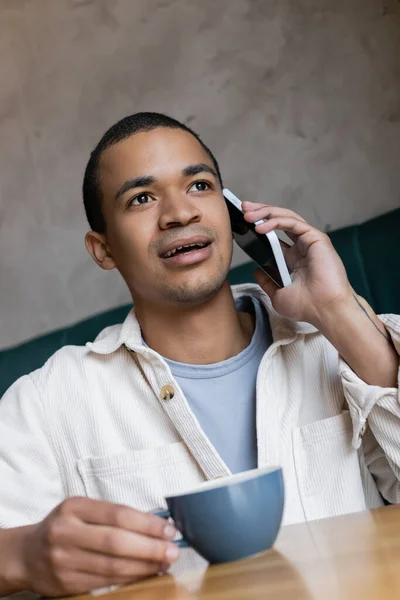 Joven afroamericano hombre teniendo llamada telefónica y sosteniendo taza de café - foto de stock