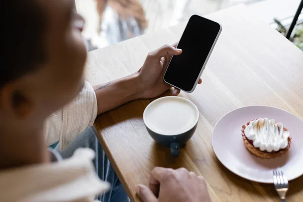 Vue grand angle de l'homme afro-américain flou tenant smartphone avec écran vide près de tasse de cappuccino — Photo de stock