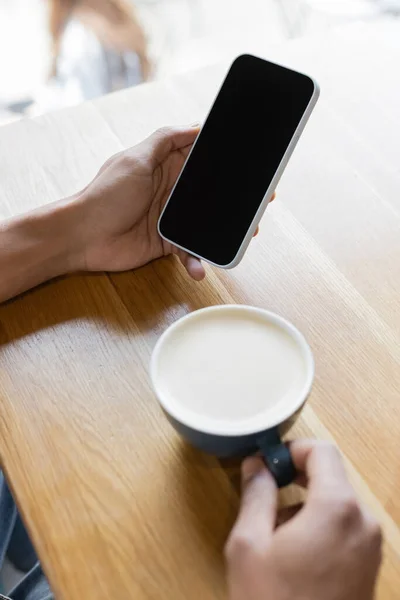 Vista cortada de homem americano africano desfocado segurando smartphone com tela em branco perto da xícara de cappuccino — Fotografia de Stock