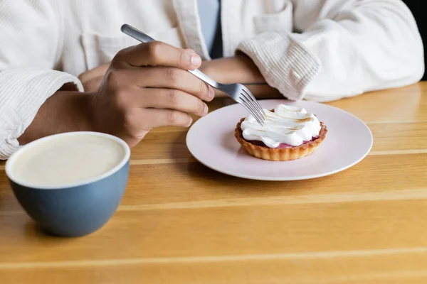 Vista cortada do homem americano africano segurando garfo perto de torta e xícara de cappuccino — Fotografia de Stock