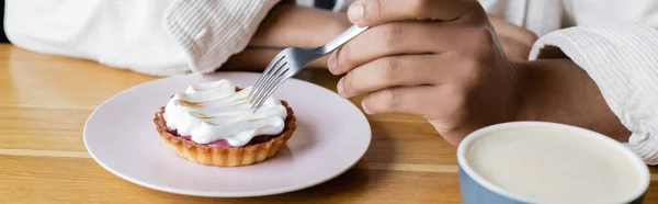 Cropped view of african american man holding fork near tart and cup of cappuccino, banner — Stock Photo