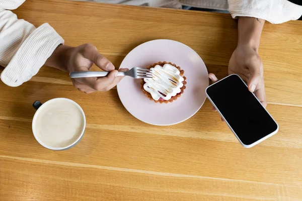 Von oben: Afrikanisch-amerikanischer Mann hält Smartphone in der Nähe von leckerer Torte und Tasse Cappuccino — Stockfoto