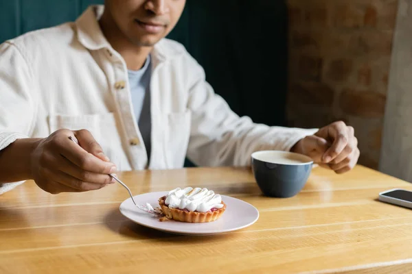 Vista ritagliata di giovane uomo afro-americano che tiene la forchetta vicino alla gustosa crostata sul piatto vicino a tazza e smartphone — Foto stock