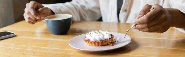 Vista recortada del joven afroamericano sosteniendo tenedor cerca de la sabrosa tarta en el plato cerca de la taza y el teléfono inteligente, pancarta - foto de stock