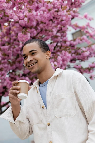 Vista basso angolo di felice uomo afro-americano in possesso di caffè per andare vicino albero di ciliegio rosa — Foto stock