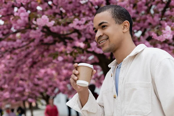 Felice uomo africano americano che tiene il caffè per avvicinarsi albero di ciliegio rosa — Foto stock