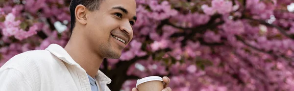 Happy african american man holding coffee to go near pink cherry tree, banner — Stock Photo