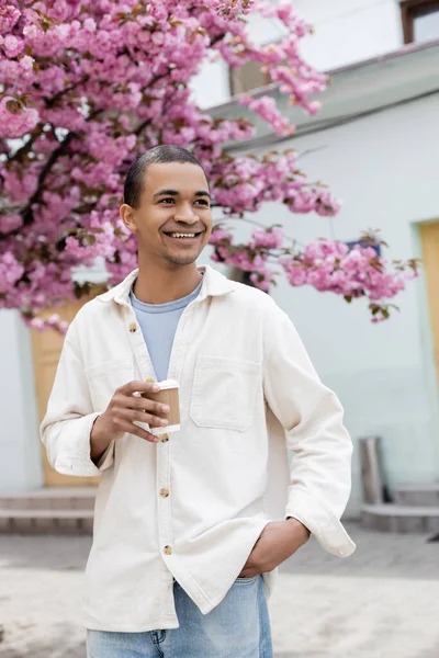 Sorrindo afro-americano homem segurando café para ir perto de cerejeira rosa na rua — Fotografia de Stock